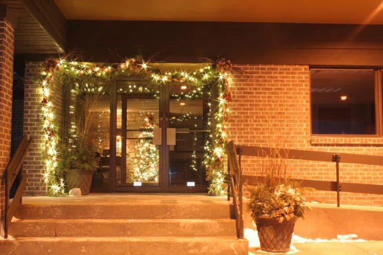The entrance way to an office building it outlined with lit greenery. A Christmas tree with white lights can be seen through the glass.
