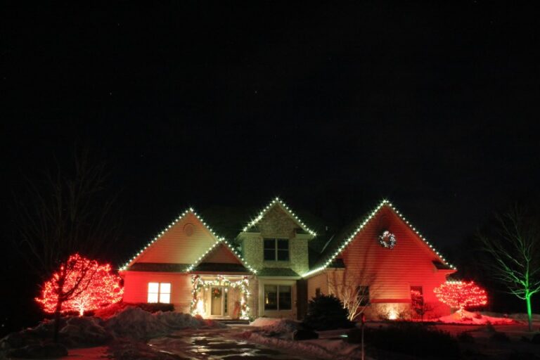 A night time view of a house with its roof outlined with white lights, lit greenery, and red and green ambient lighting.