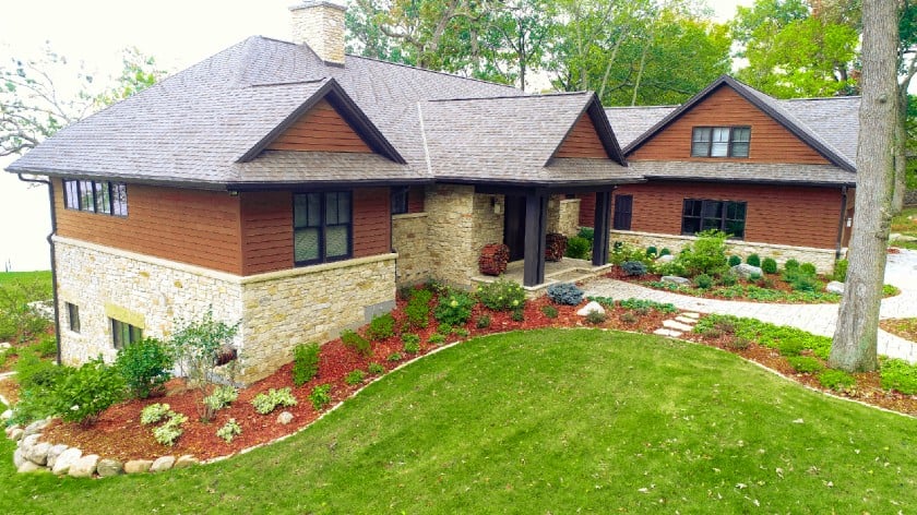 Light and dark green shrubs and bushes line the front and front walk of a house. The house is one-story and has brown siding with light brown stone along the bottom.